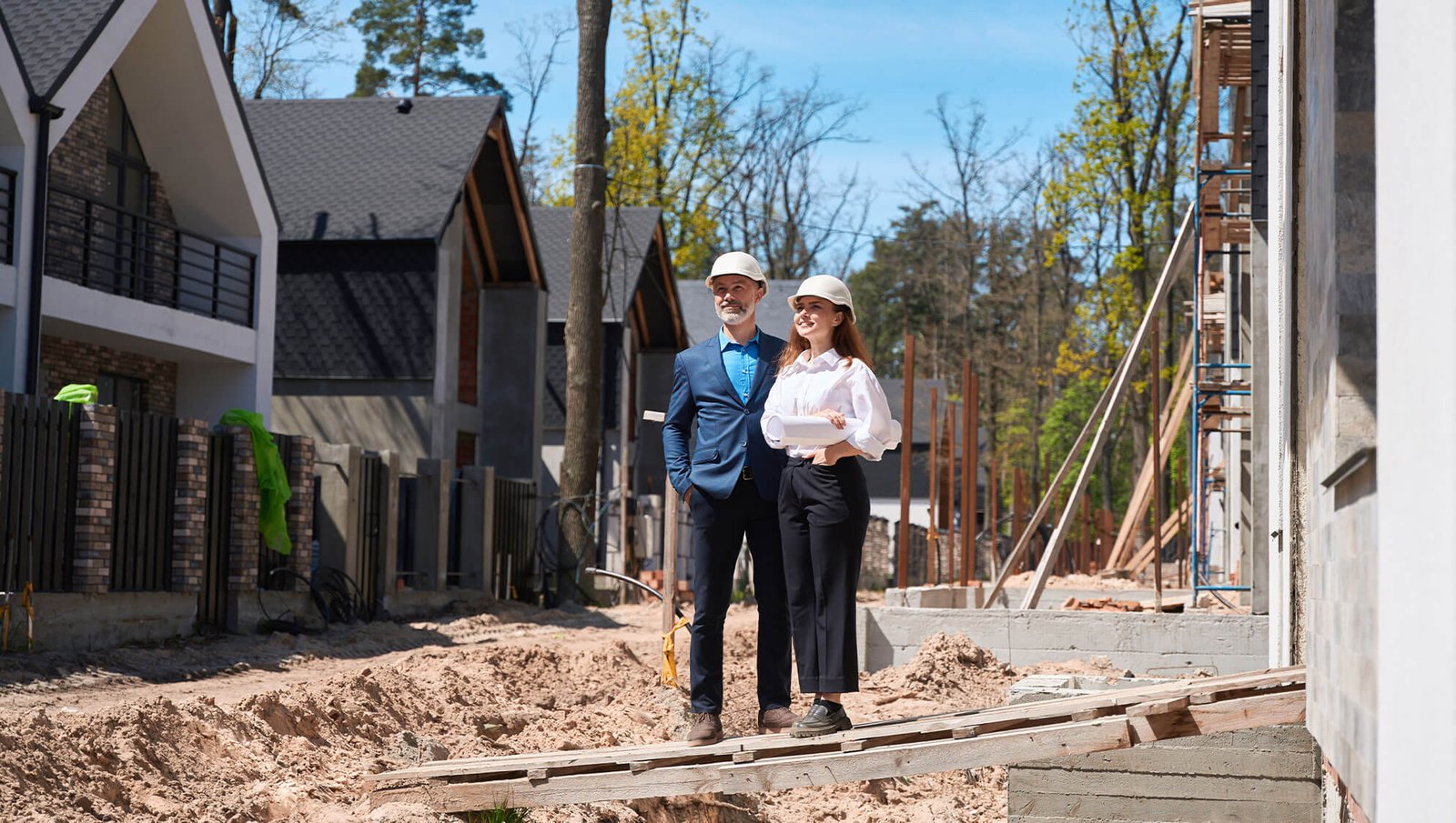 A male and female architect wearing hard hats stand on a construction site, holding blueprints, with modern houses under construction nearby.