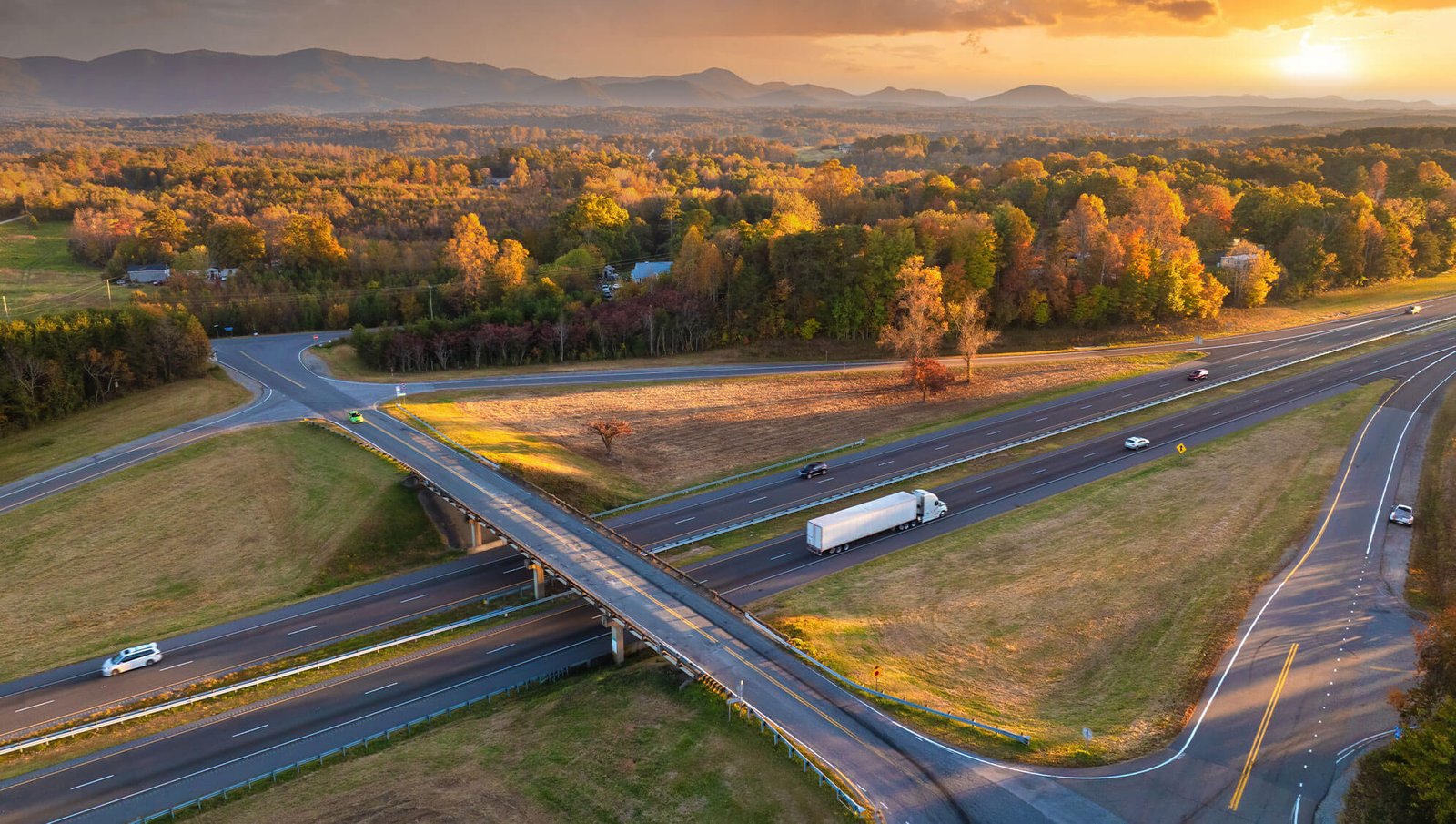 An aerial view of a highway intersection surrounded by vibrant autumn foliage, rolling hills, and a golden sunset in the background.