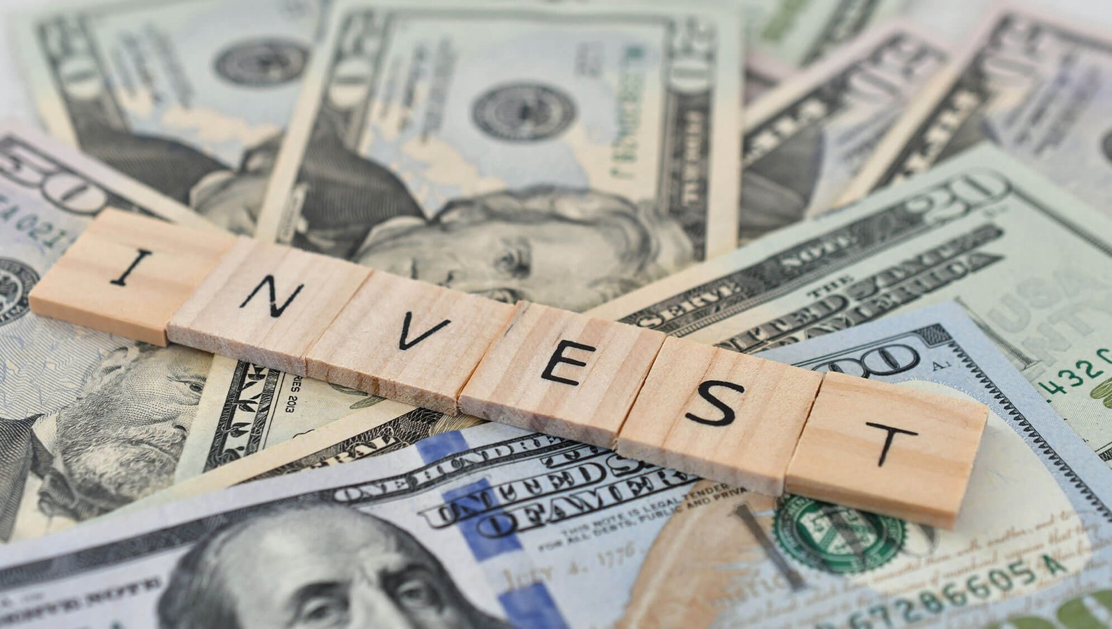 Wooden tiles spelling "INVEST" are placed on a scattered background of U.S. dollar bills, symbolizing financial growth and investment.