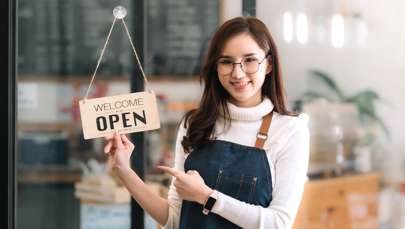 A smiling woman wearing glasses and an apron holds a "Welcome, We Are Open" sign at the entrance of a bright and cozy shop or cafe.