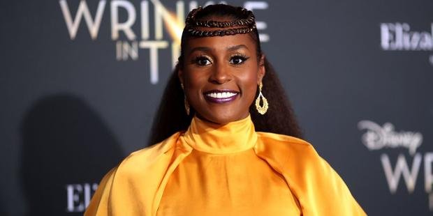 Issa Rae’s vibrant yellow outfit with elegant gold earrings and braided hair smiles at a premiere event, standing in front of a branded backdrop