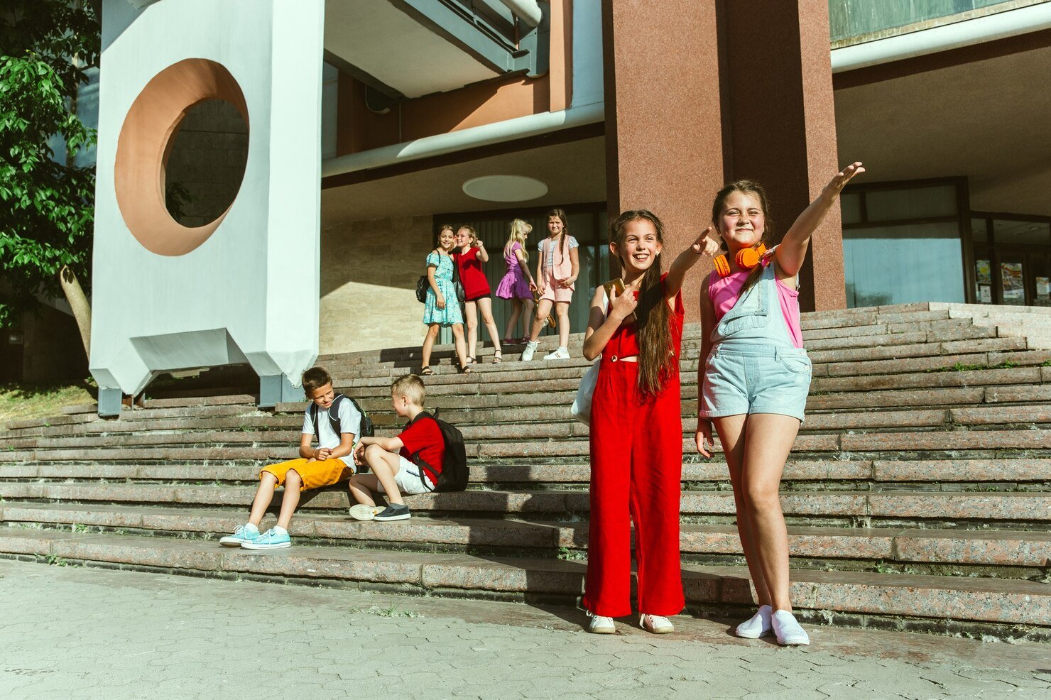 A group of cheerful children gather on the steps of a modern building, chatting, smiling, and enjoying a sunny day in a casual outdoor setting.