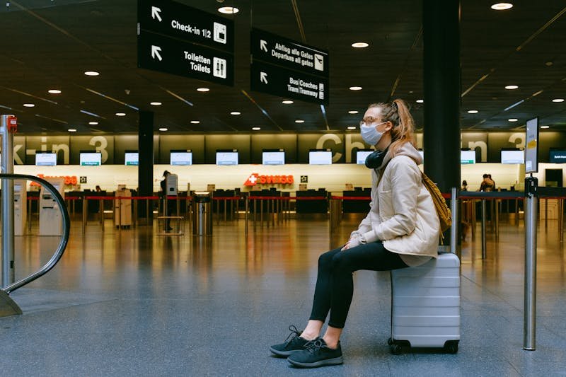 A traveler girl wearing a face mask sits on a suitcase in an airport terminal, and waiting for flight